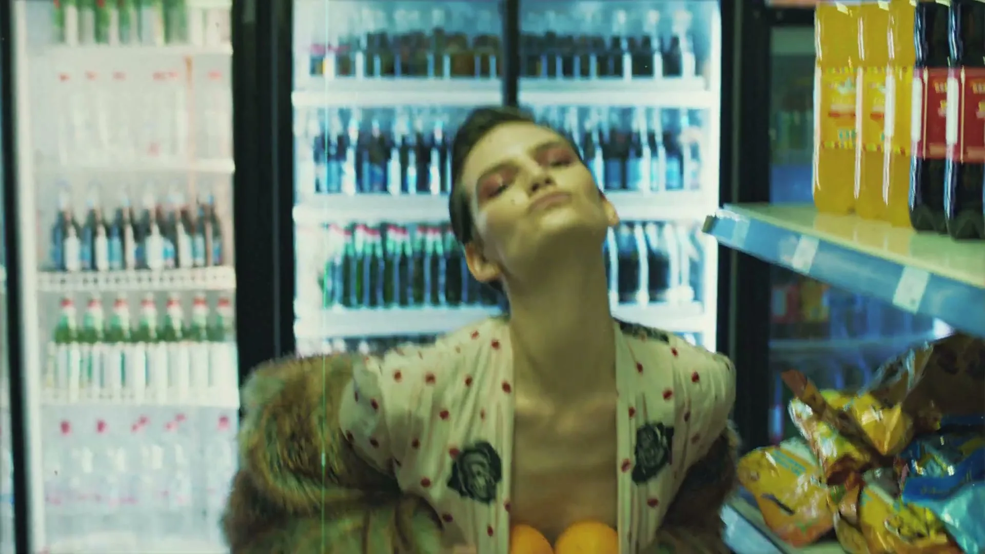 A young woman with short hair and a fur coat holds two oranges to her chest in a convenience store aisle, looking up and smiling.