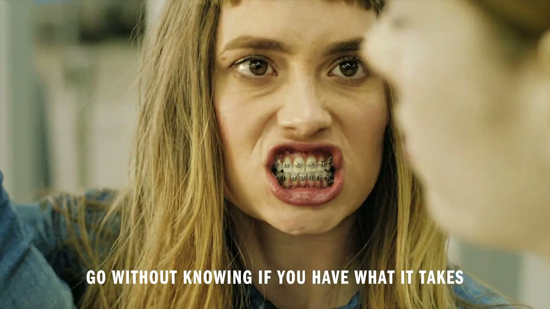 A young woman with braces and bangs looks at her reflection in a mirror, making an angry face.