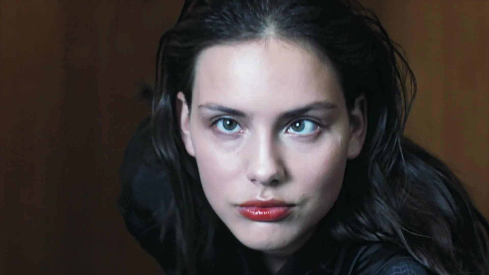A young woman with dark hair and red lipstick looks directly at the camera, her eyes slightly crossed, in a close-up shot.