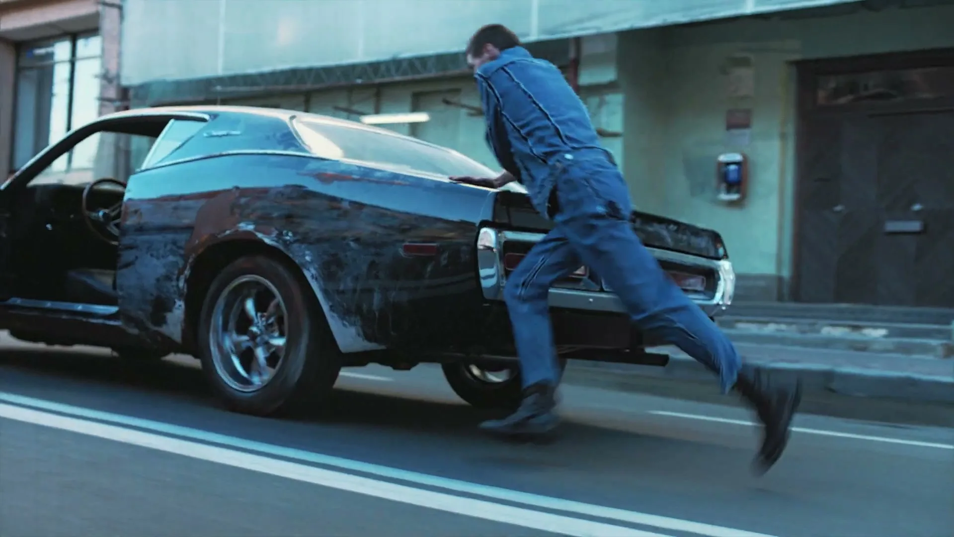 A young man in a denim outfit pushes a beat-up black car down a city street during the day.