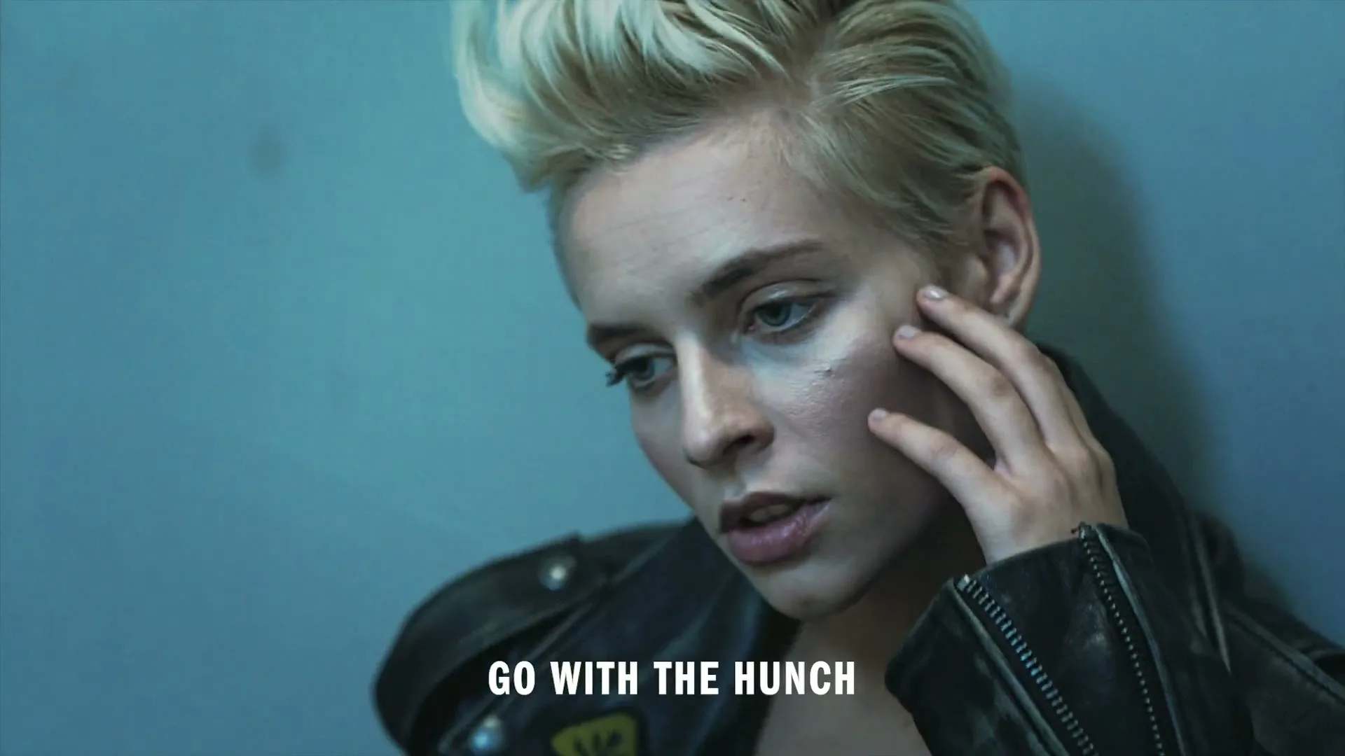 A young woman with short blonde hair and a leather jacket looks down, touching her face, against a blue wall.