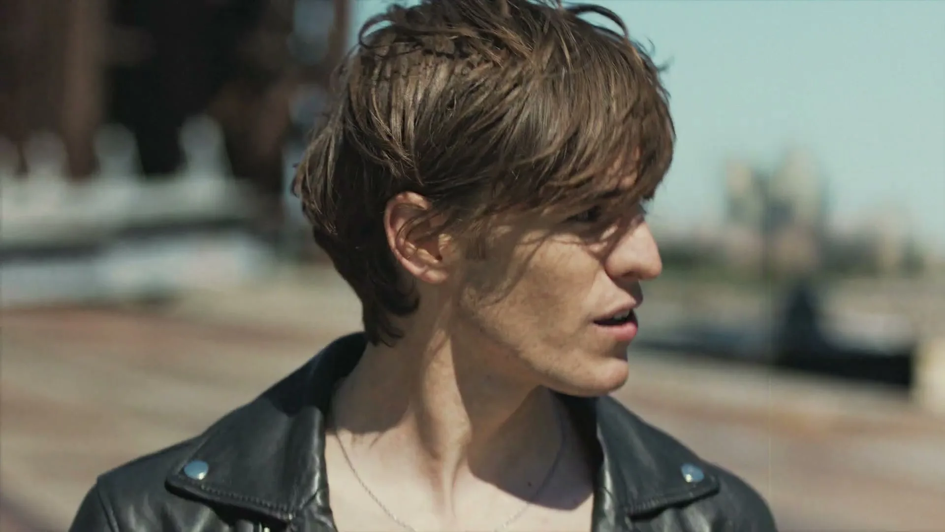 A young man with brown hair and a black leather jacket looks around on a sunny rooftop.