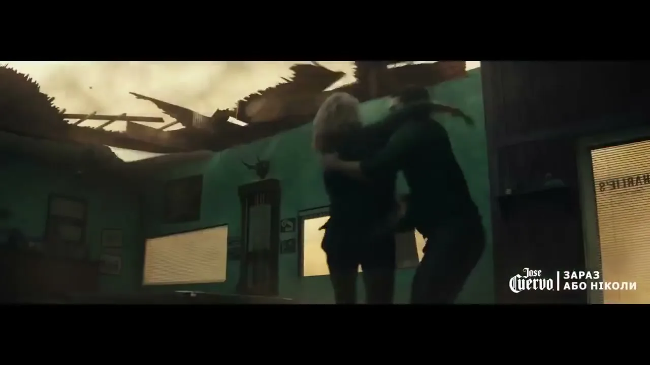 A man and woman embrace in a destroyed room with a collapsed roof, as debris falls around them, suggesting a chaotic environment.