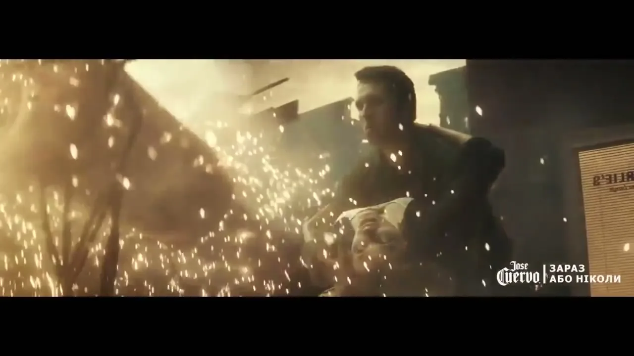 A man in a dark shirt carries a woman in a white dress through a shower of sparks and debris in a collapsing building.