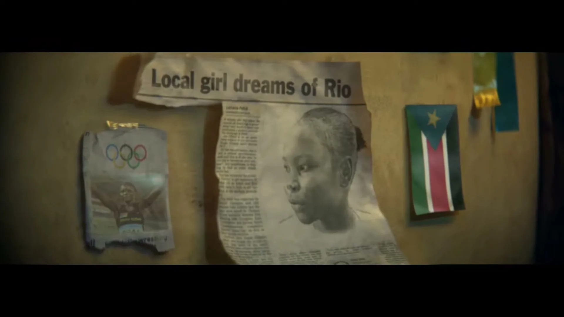 A newspaper clipping with the headline "Local girl dreams of Rio" and a black and white photo of a young girl is taped to a wall, next to a small flag.