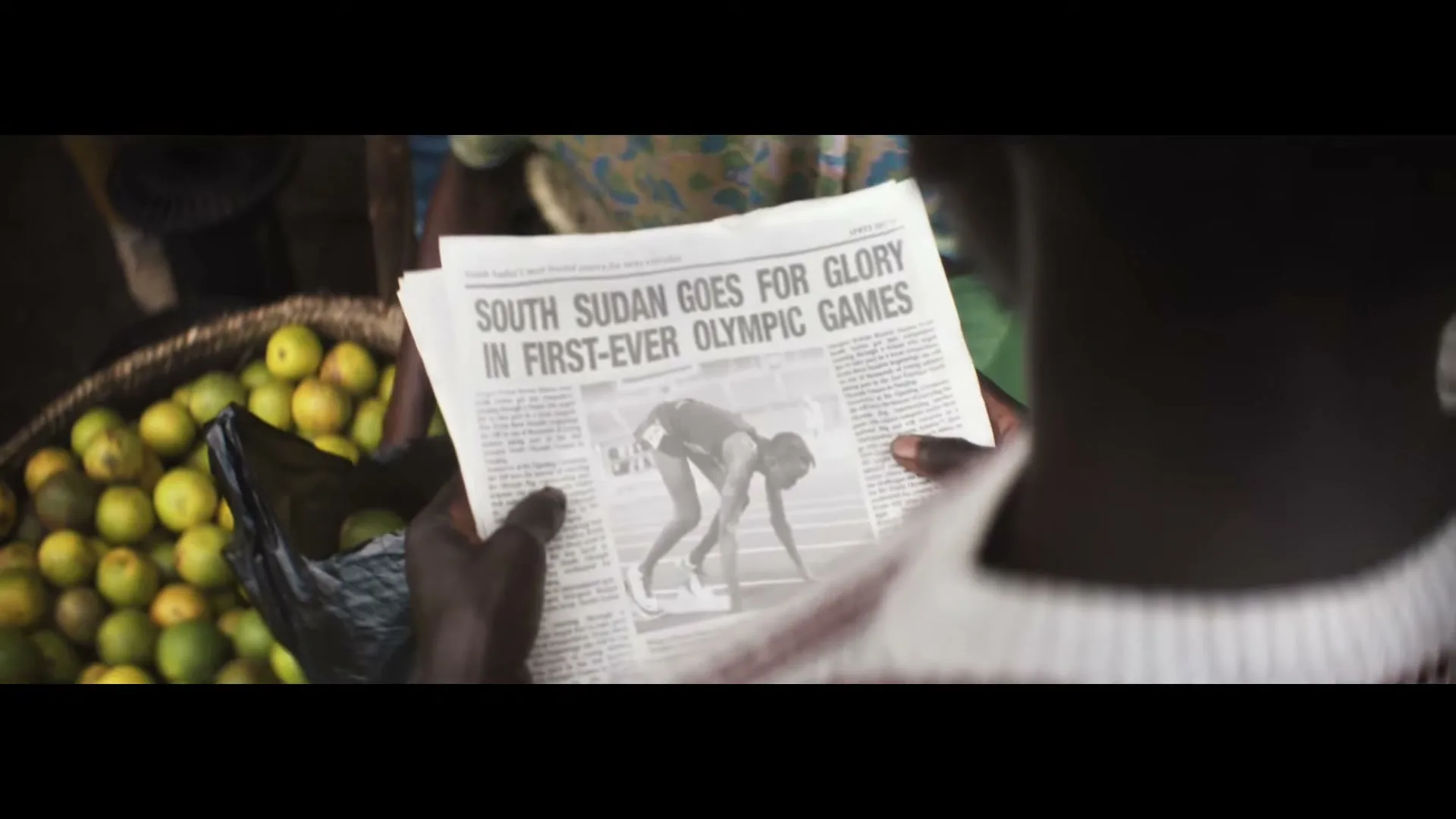 A person's hands hold a newspaper with the headline "SOUTH SUDAN GOES FOR GLORY IN FIRST-EVER OLYMPIC GAMES" over a basket of fruit in a market.