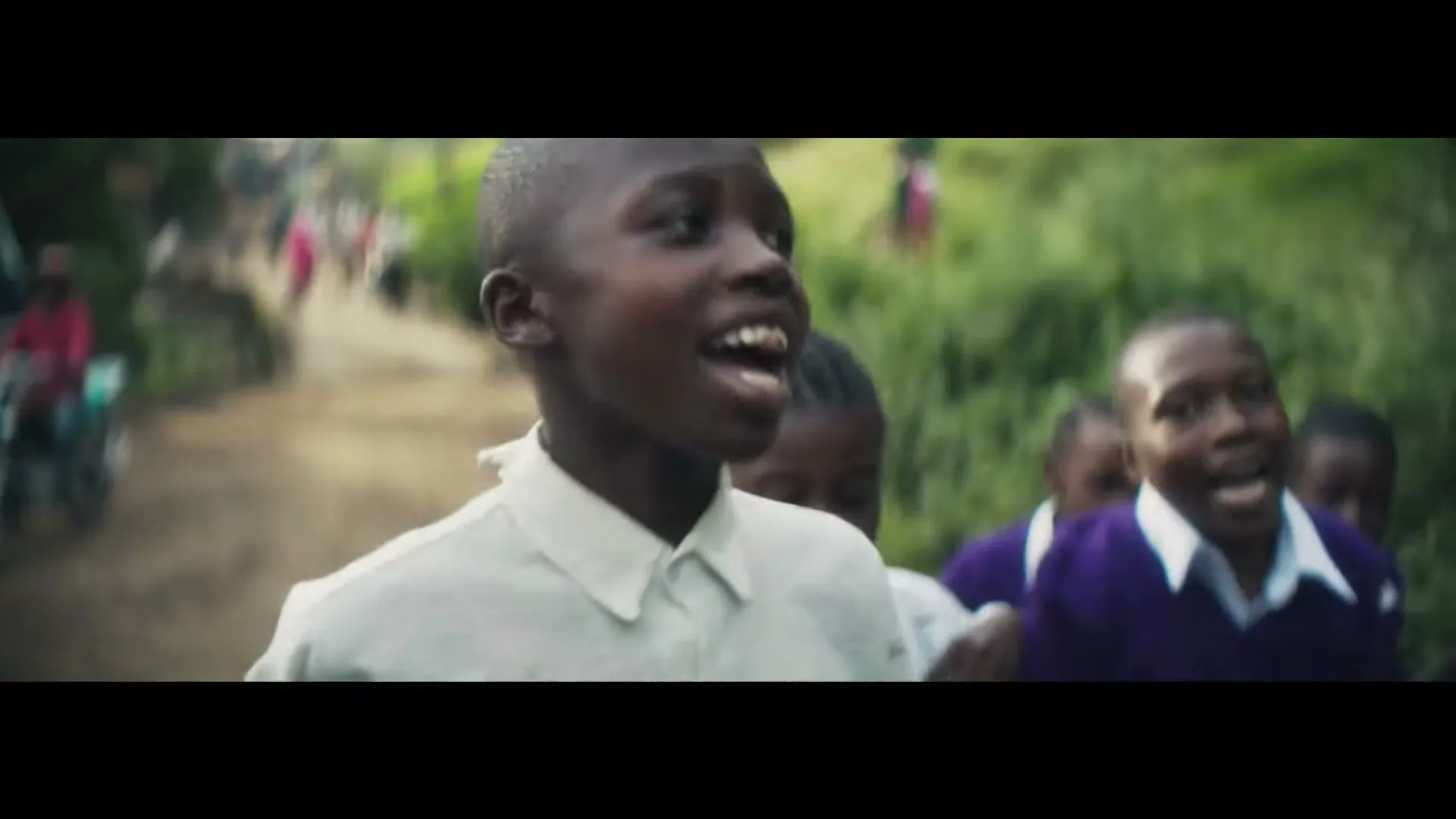 A group of African children, some in school uniforms, run excitedly down a dirt road, cheering and smiling in a rural setting.