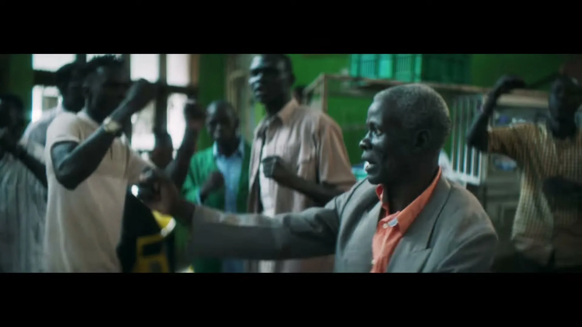 An older man with dark skin and grey hair, wearing a grey suit jacket and orange shirt, sings and gestures with his hands among a group of men in an interior setting.