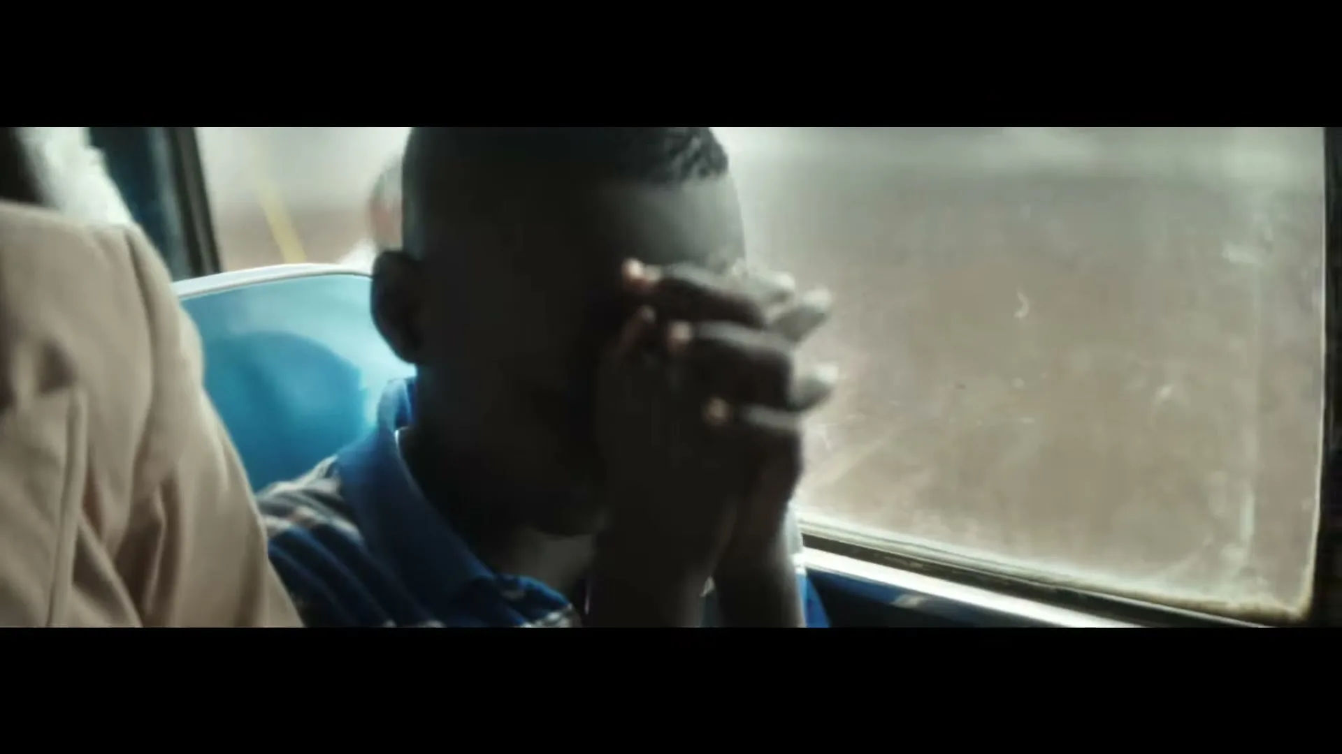 A young boy with dark skin, wearing a blue and white striped shirt, sits on a bus with his hands clasped in front of his face, looking anxious.