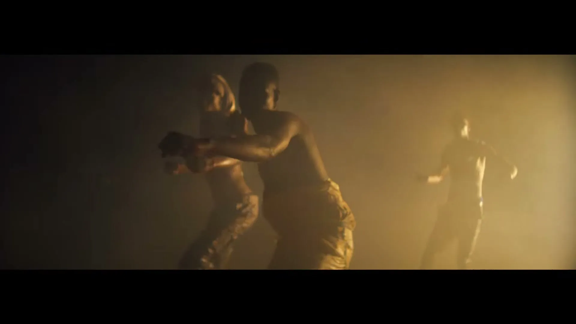 Three male athletes, shirtless and wearing athletic shorts, train in a smoky, dimly lit room, performing martial arts movements.