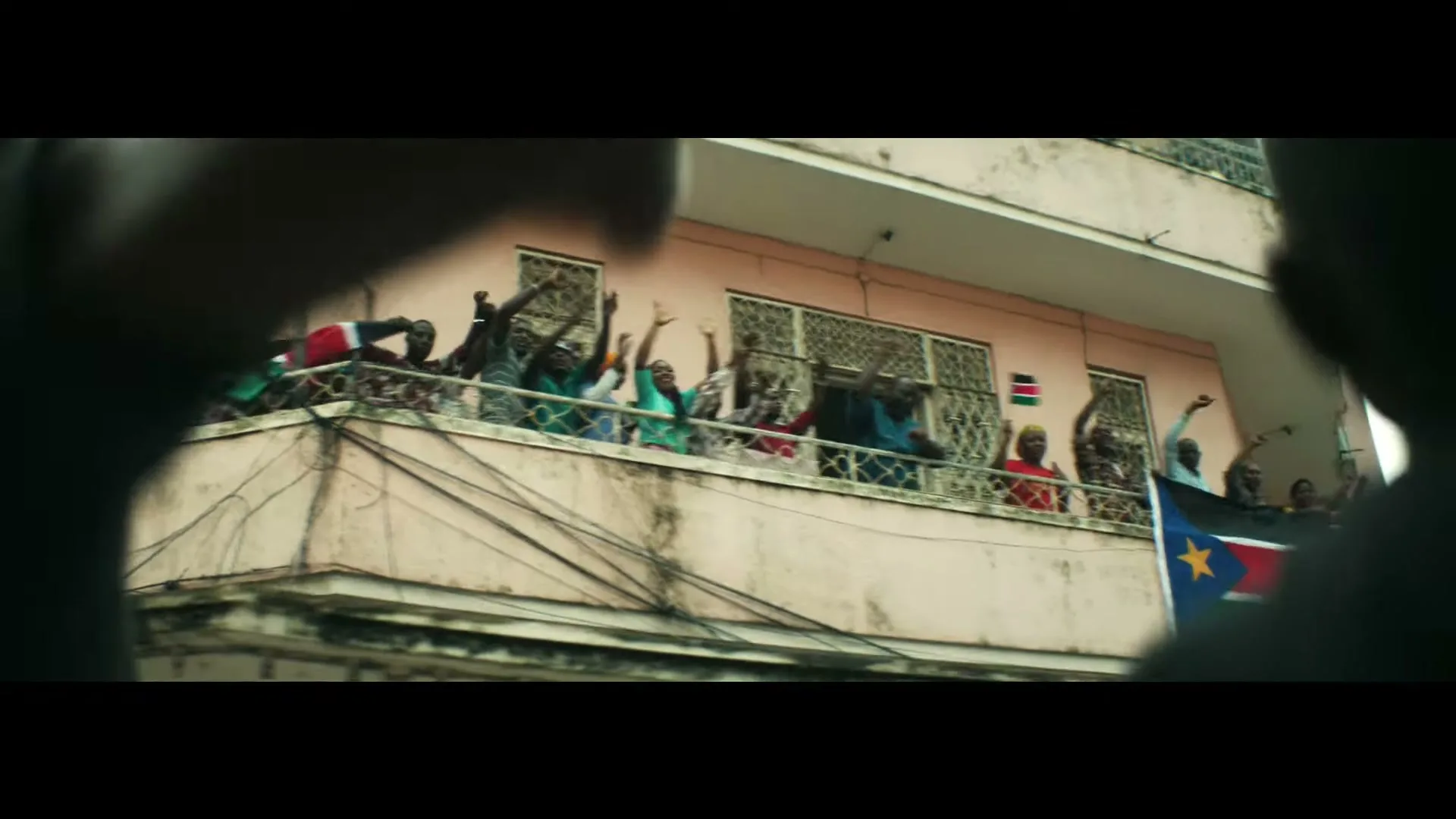 A crowd of people, mostly adults, cheer and wave their hands from a balcony of a building, with a flag hanging below.