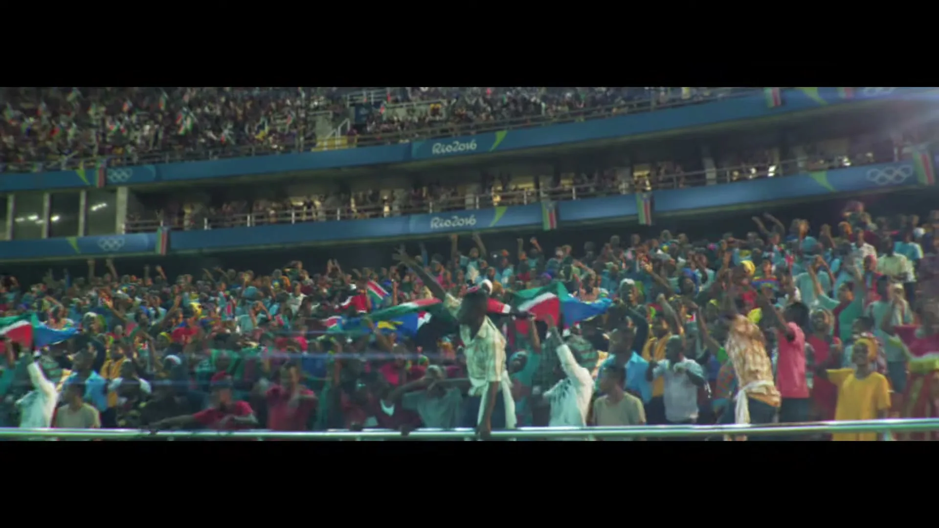 A large crowd of diverse people, many holding flags, cheer enthusiastically in a brightly lit stadium during a sporting event.