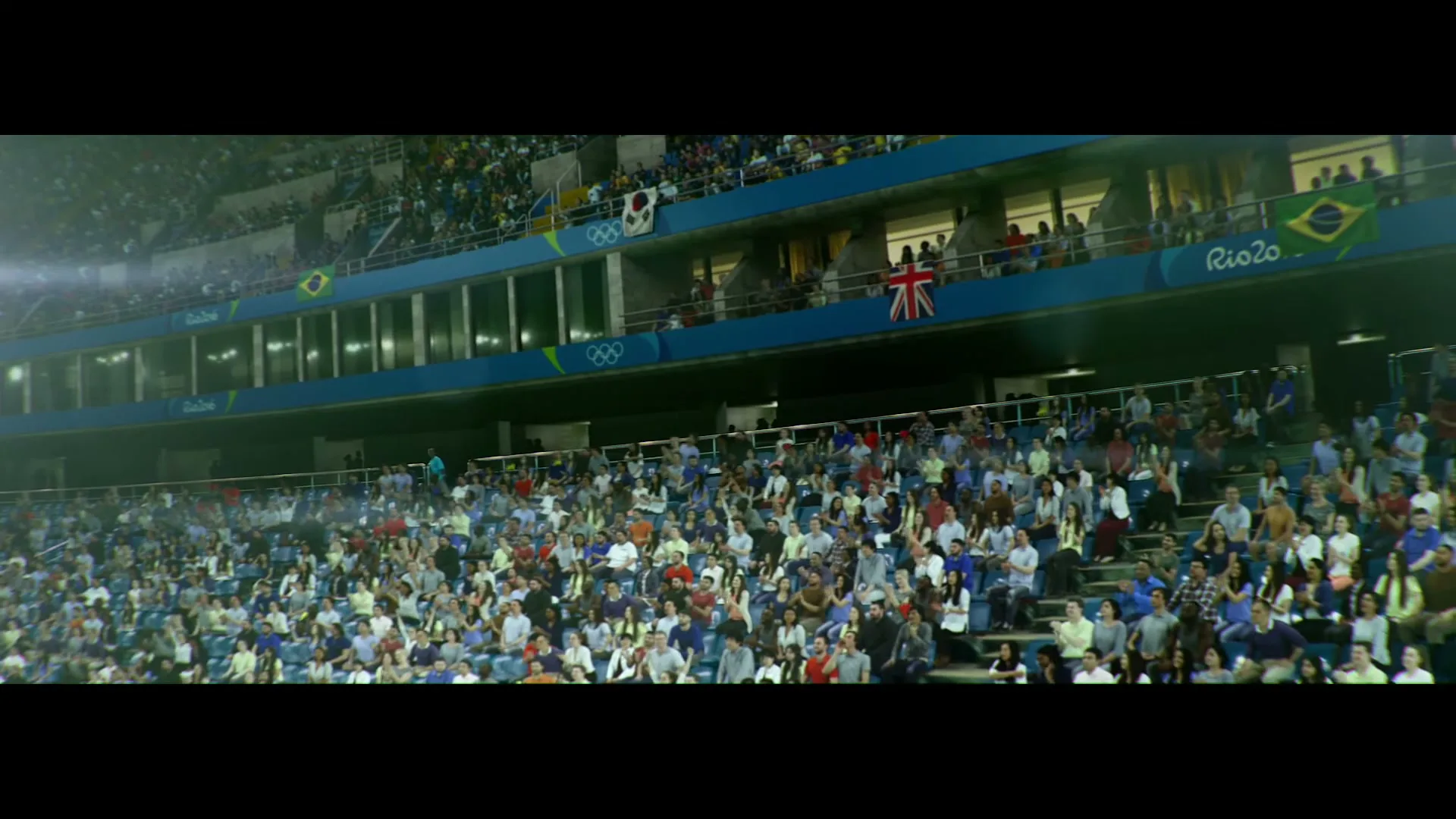 A wide shot of a stadium filled with a diverse crowd of people, with flags and Olympic rings visible in the background.