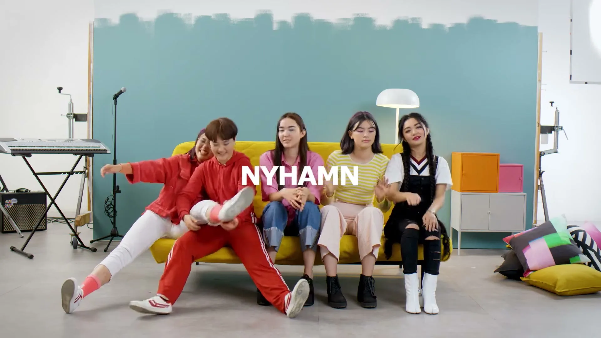 Five young women sit on a yellow couch in a studio, with one woman playfully kicking her leg up, as the word "NYHAMN" appears on screen.