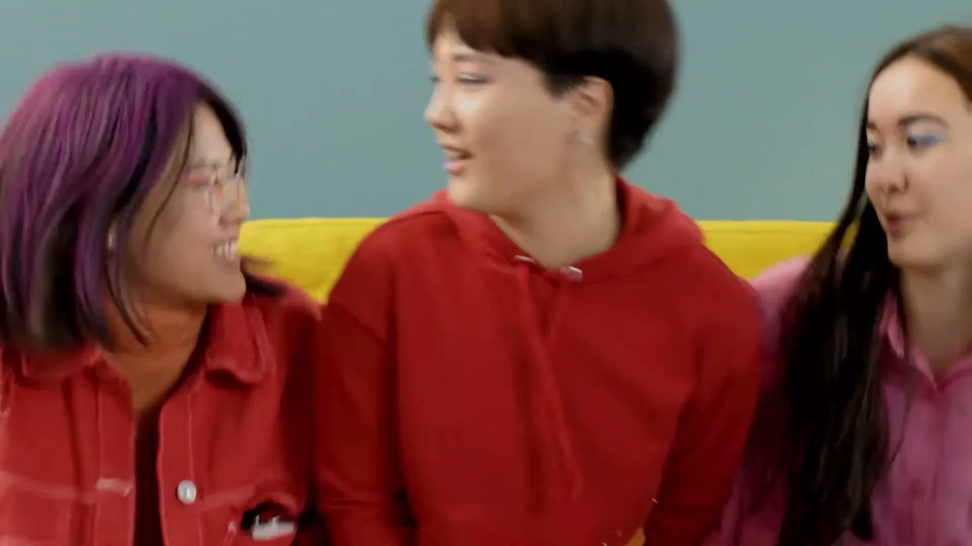 Three young women, one with purple hair, one with short brown hair, and one with long dark hair, are sitting on a yellow couch, talking and laughing.