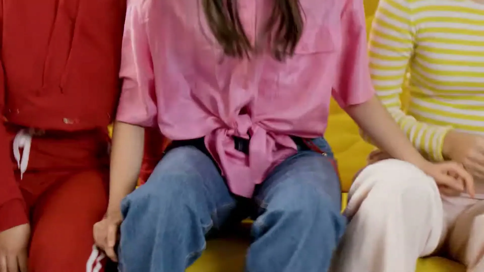 Three young women, dressed in casual clothes, are sitting closely together on a yellow couch, holding hands, in an interior setting.