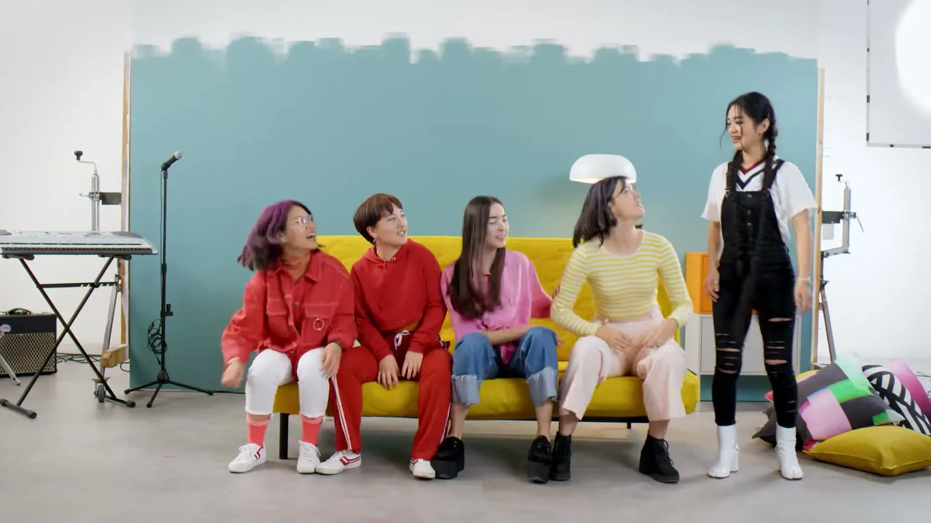 Five young women in colorful outfits are sitting and standing on a yellow couch in a studio with a blue and white wall.