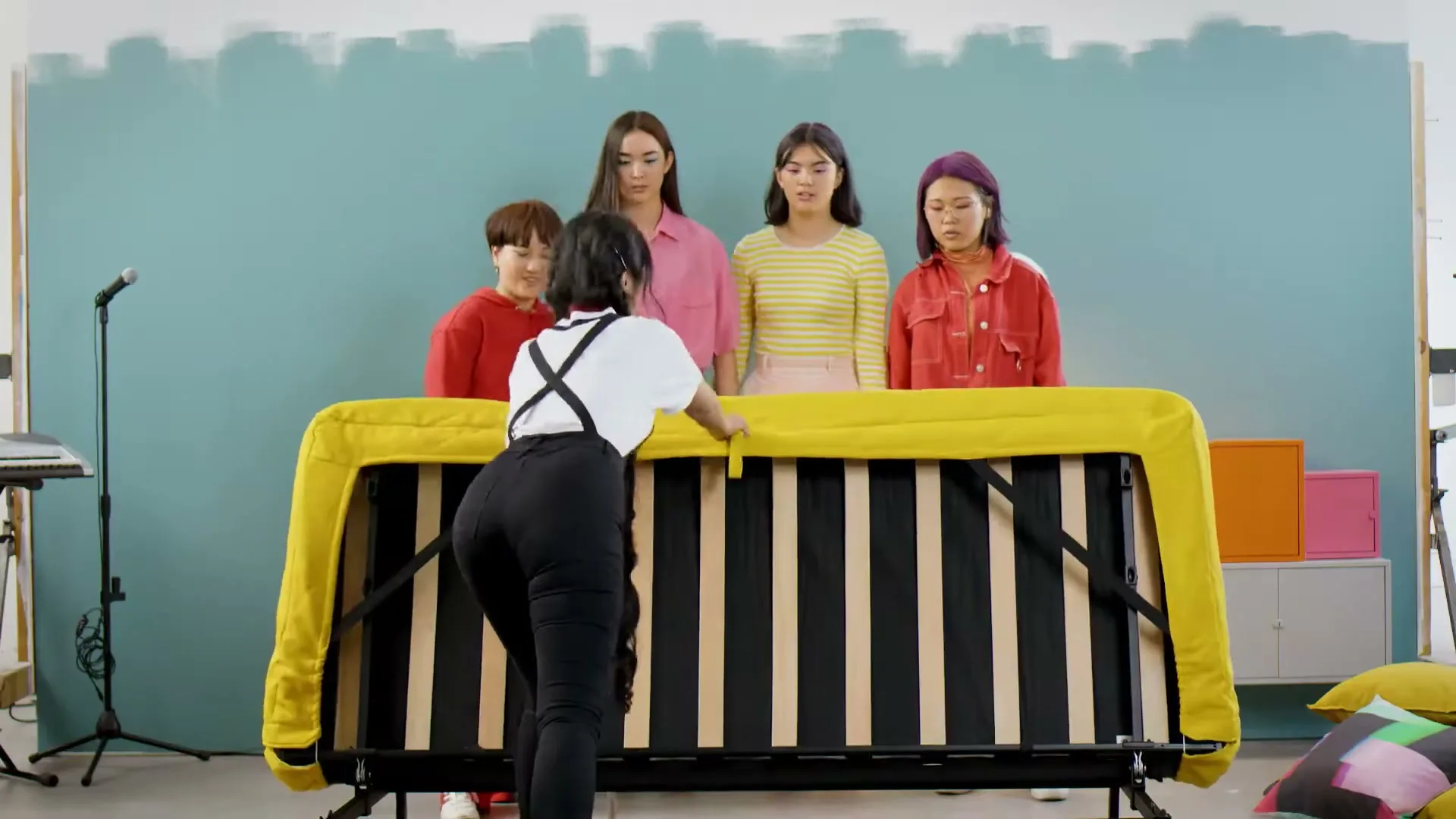 A young woman in overalls lifts a yellow sofa bed, revealing its slatted base, while four other young women watch in a brightly lit studio.