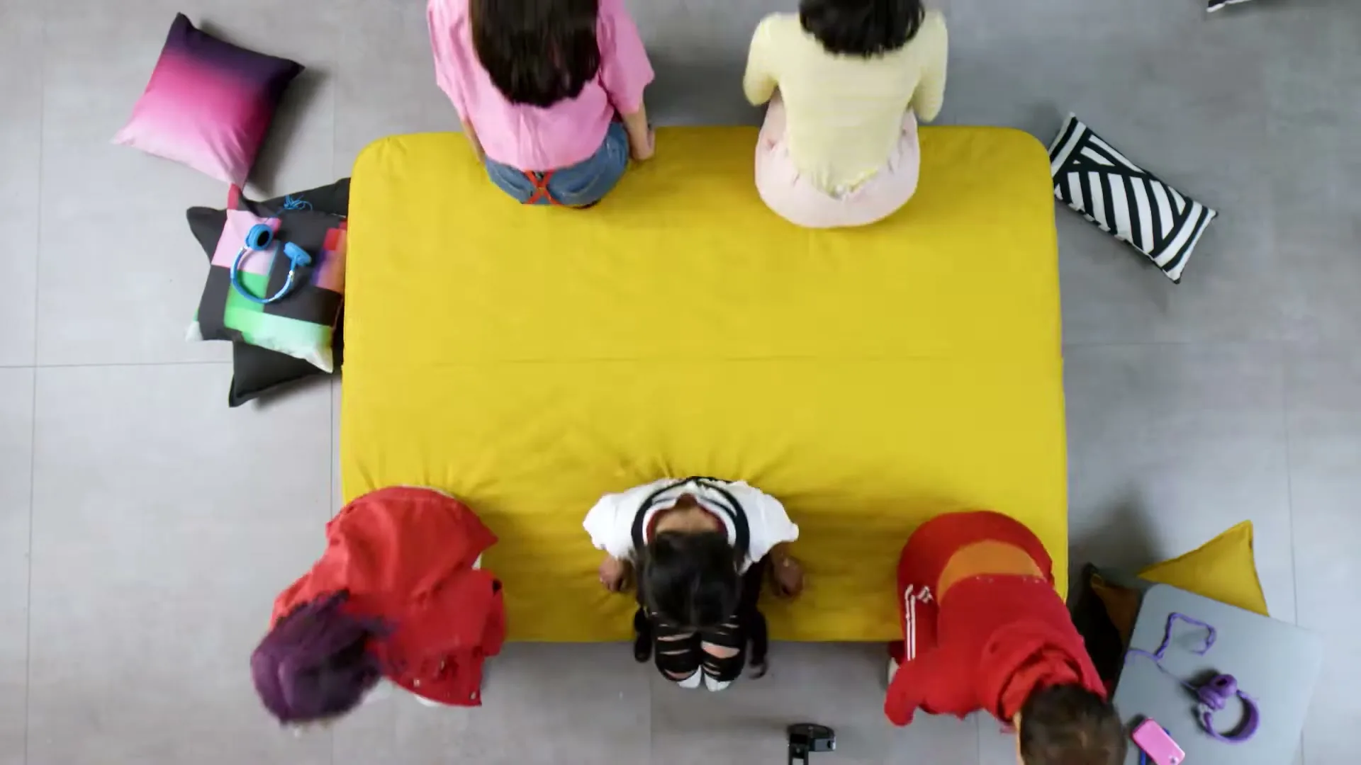 An overhead shot shows four children playing on a yellow mattress on the floor, with pillows and headphones scattered around them.