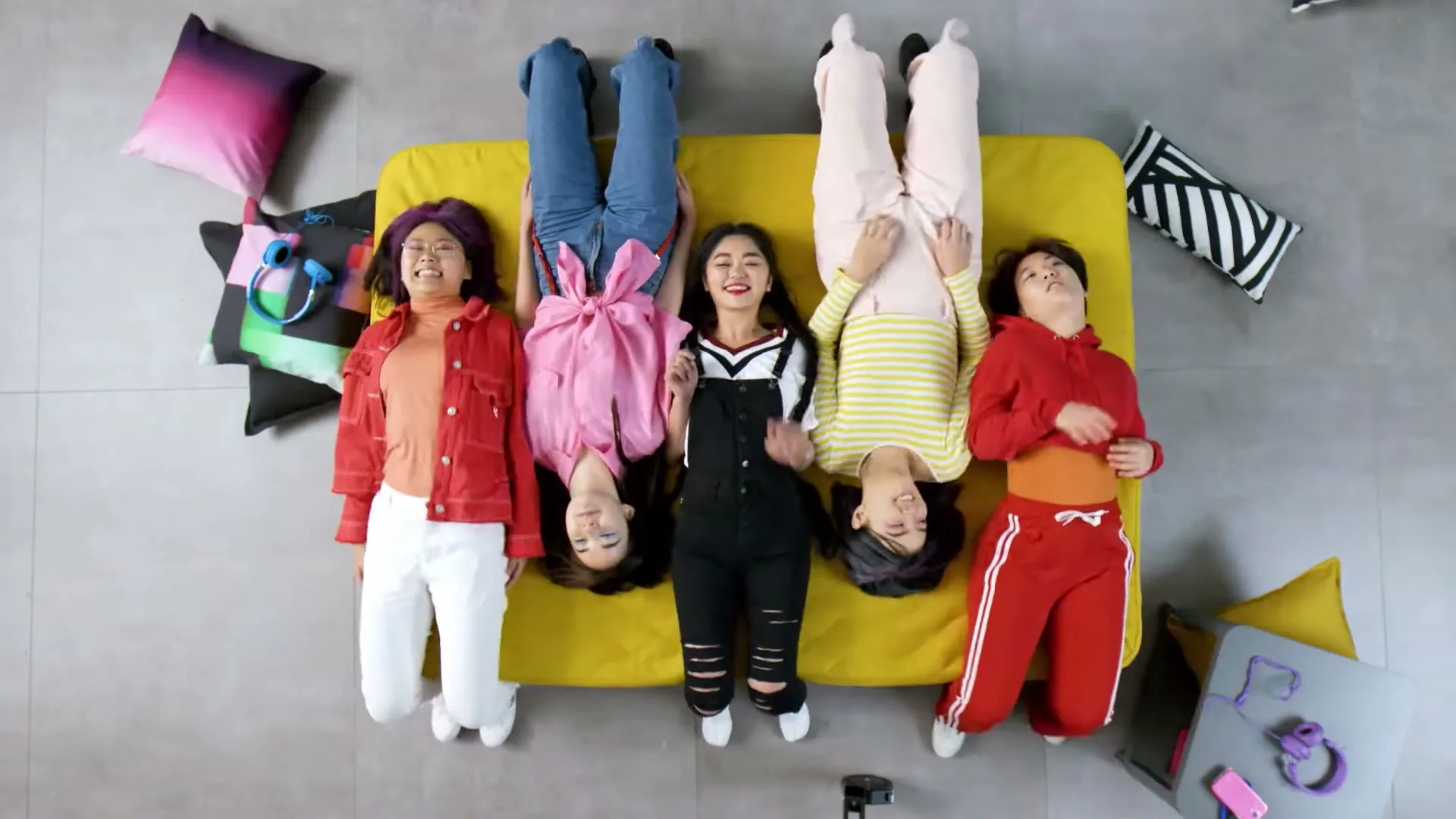 Four young women lie upside down on a yellow sofa, smiling and laughing, in a bright, modern room.