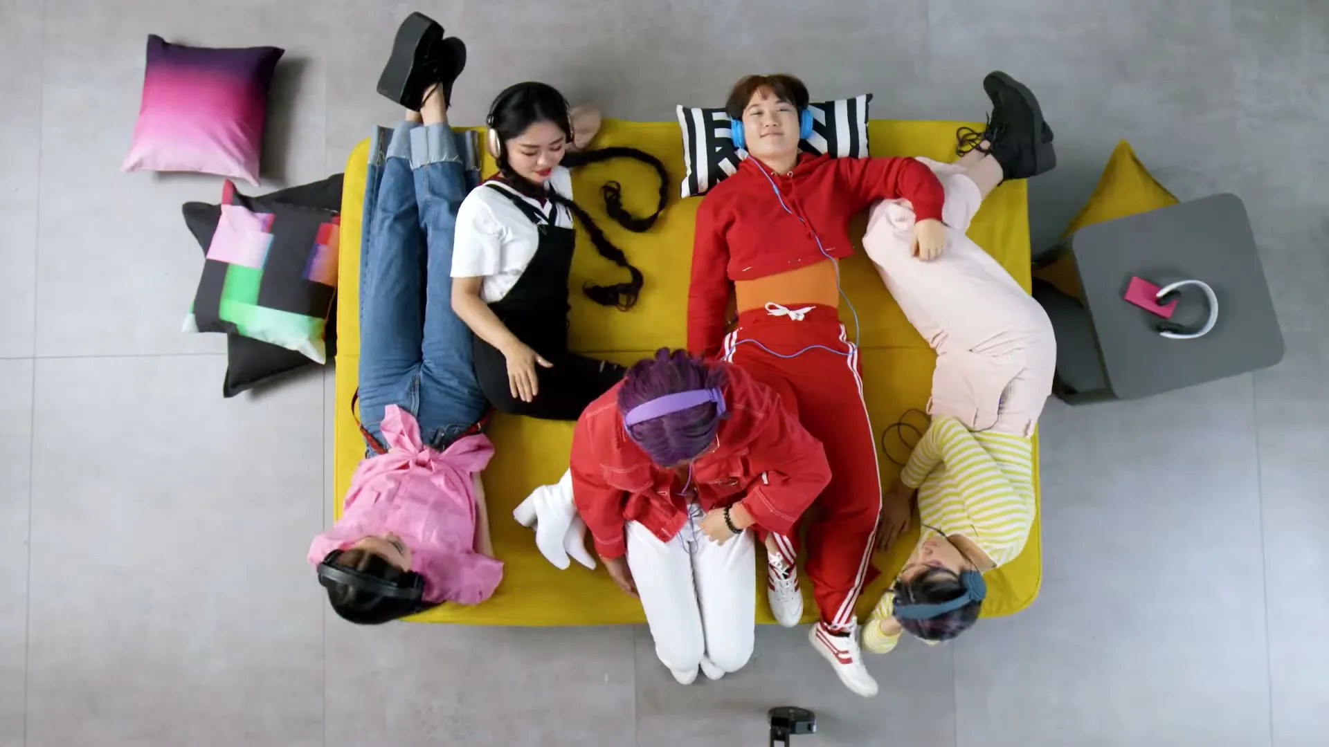 Five young people in colorful clothes and headphones lie on a yellow sofa, arranged in a star shape, seen from directly above.
