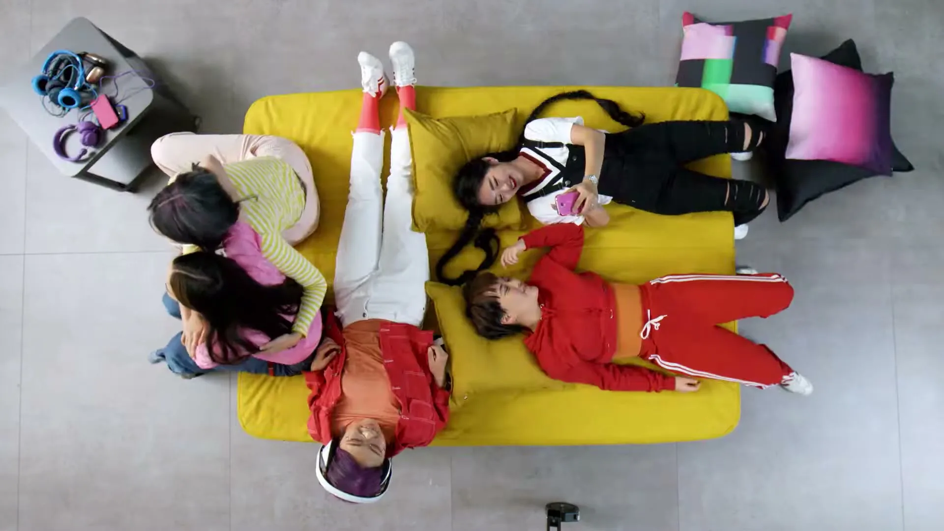 Five young people are lying on a yellow sofa and the floor around it, seen from an overhead top-down angle in a modern living room.