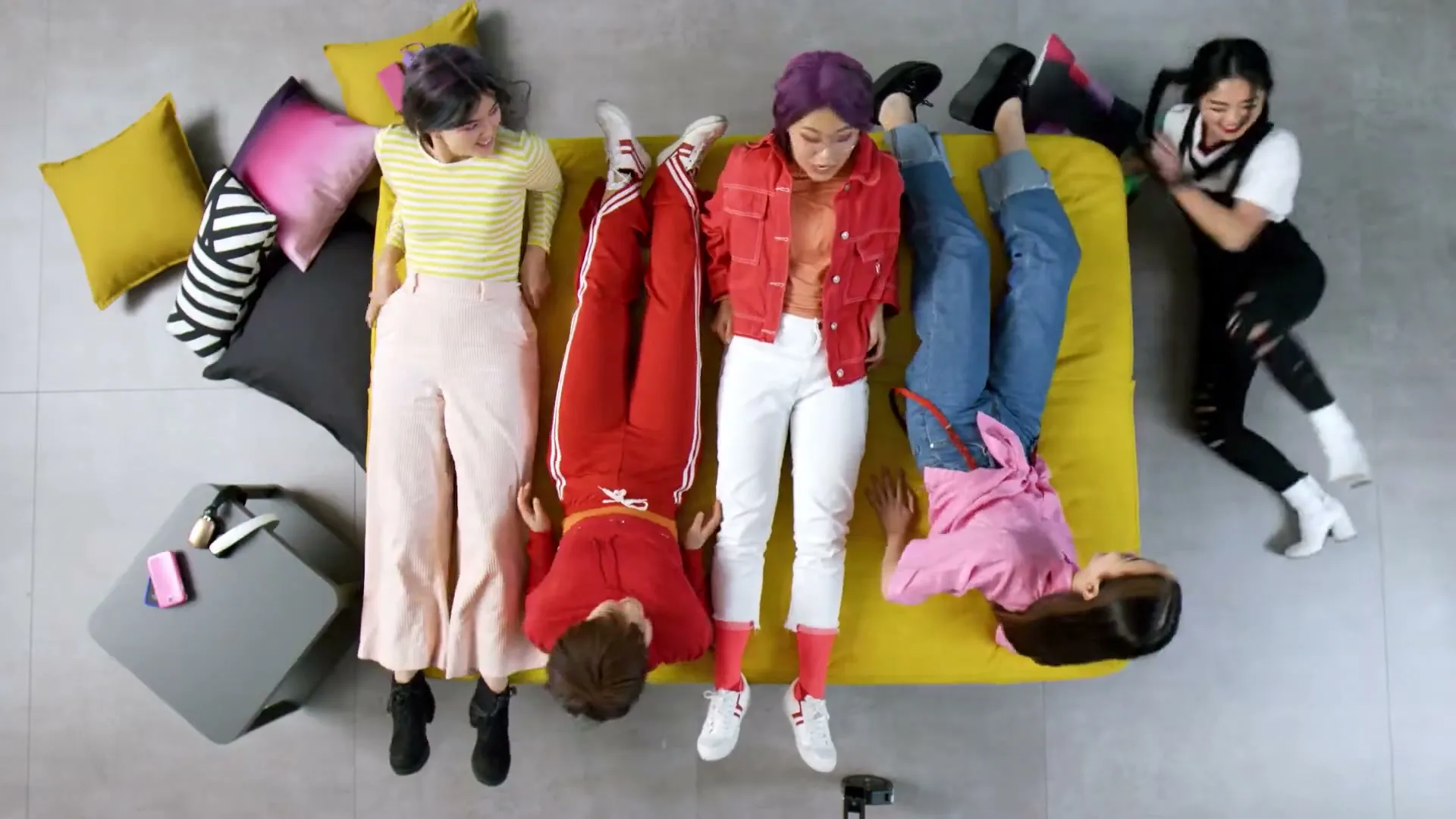 Four young women with colorful hair lie on a yellow sofa, with one woman on the right playfully kicking her legs up in a bright, modern living room.