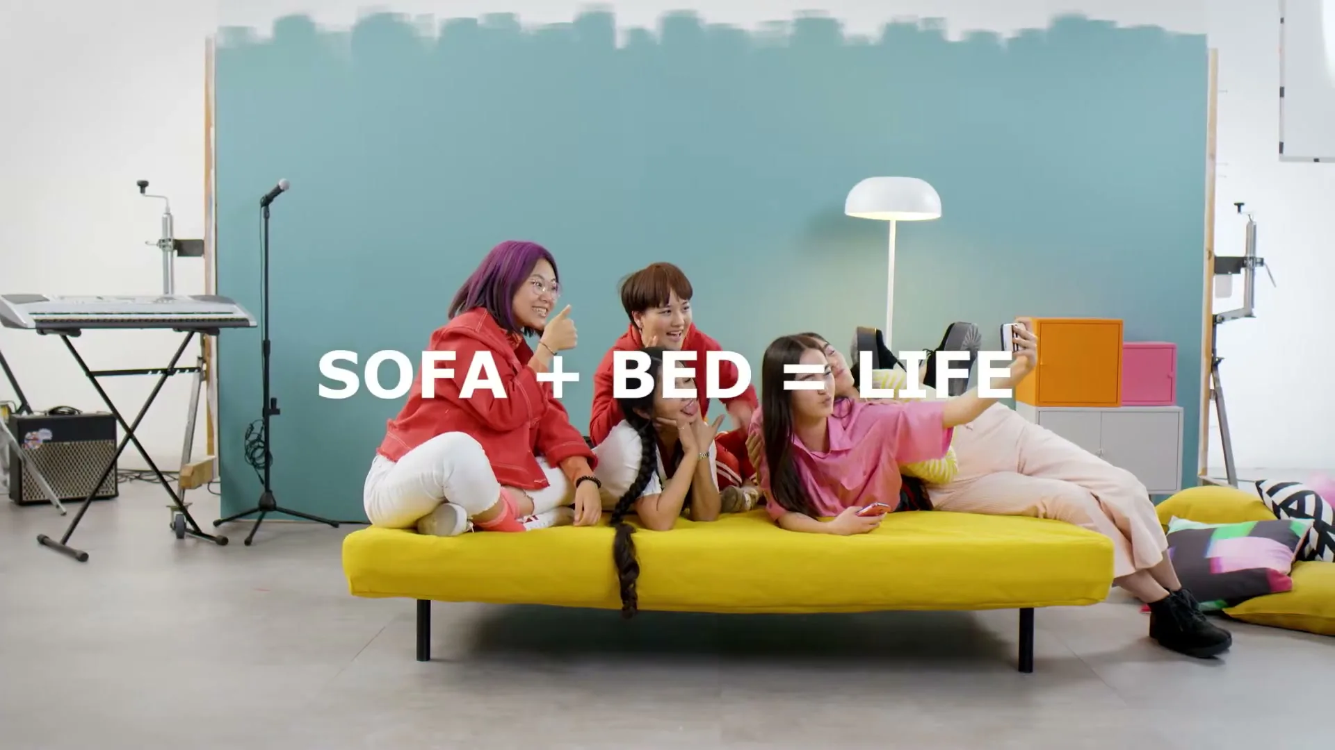 A group of five young adults are taking a selfie on a yellow sofa bed in a studio with a blue and white wall.