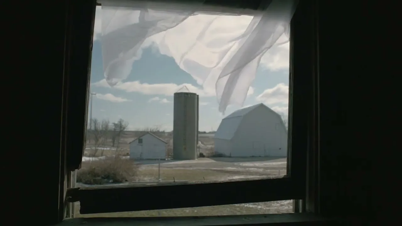 A white curtain blows in the wind through an open window, revealing a snowy rural landscape with a white barn and silo under a blue sky.