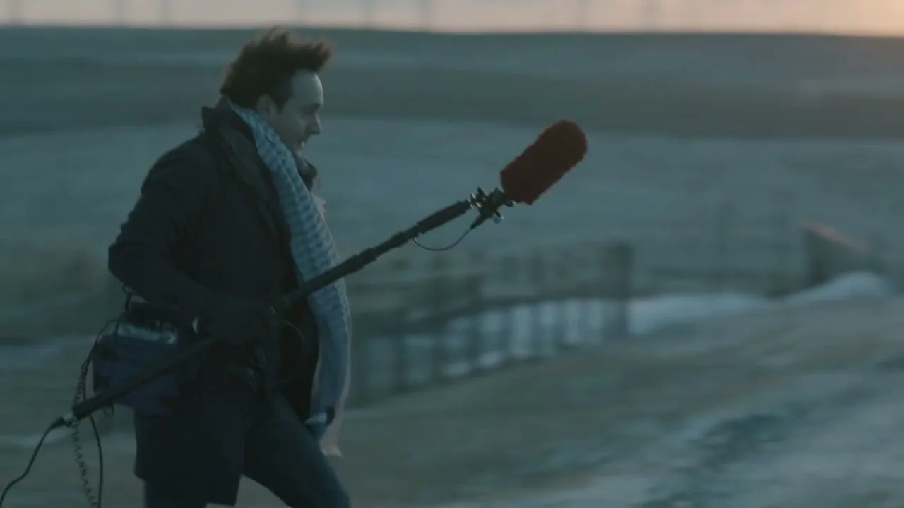 A male sound engineer with a boom mic walks across a desolate, snowy landscape at sunrise, capturing ambient sounds.