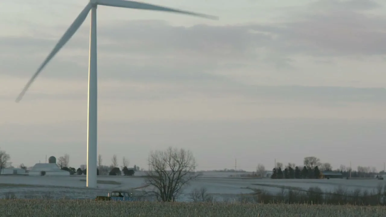 A large white wind turbine with spinning blades stands tall in a vast, snow-dusted field under a cloudy sky, with a small blue truck visible in the distance.