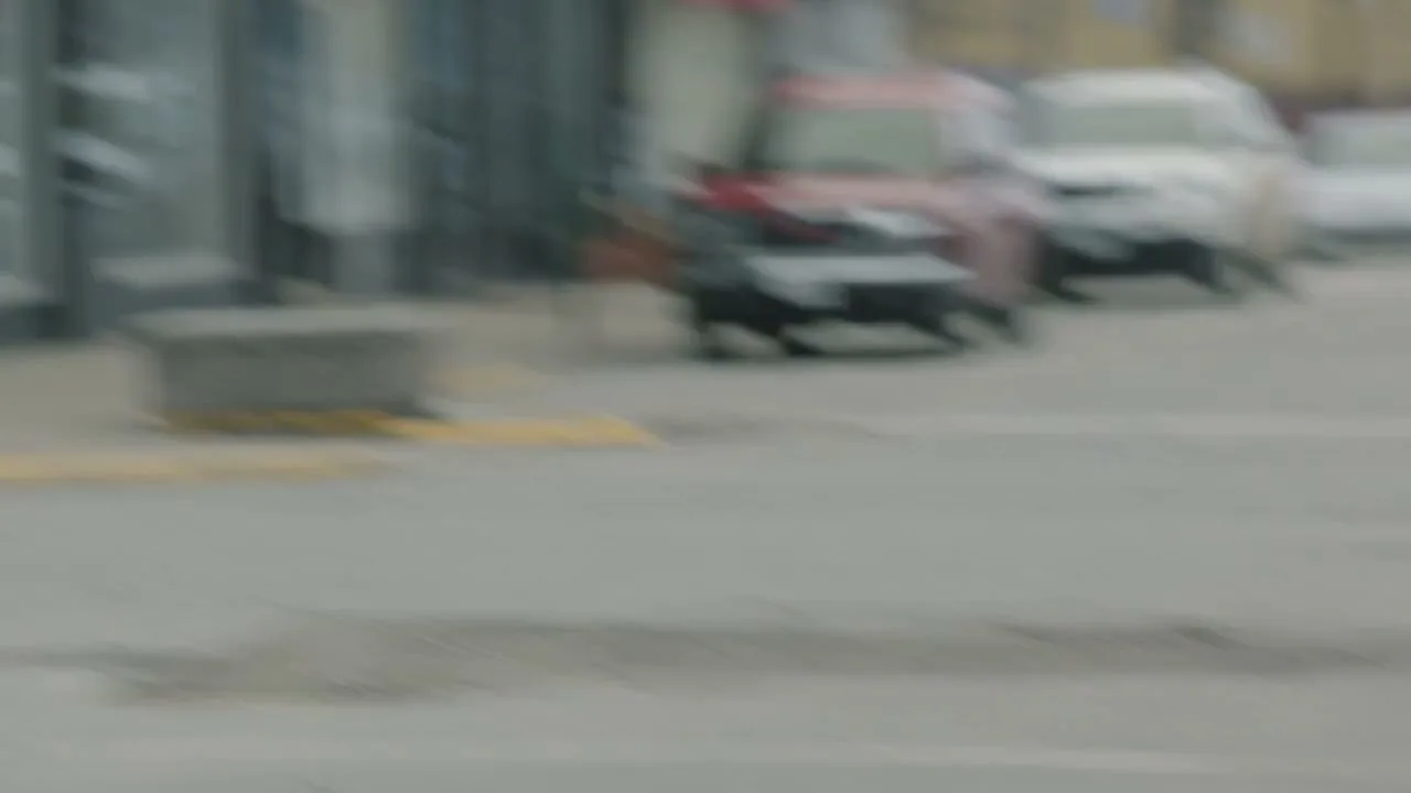 A woman in a light jacket and dark pants runs across a street, chasing a hat that is rolling away in the wind, with blurred buildings and cars in the background.