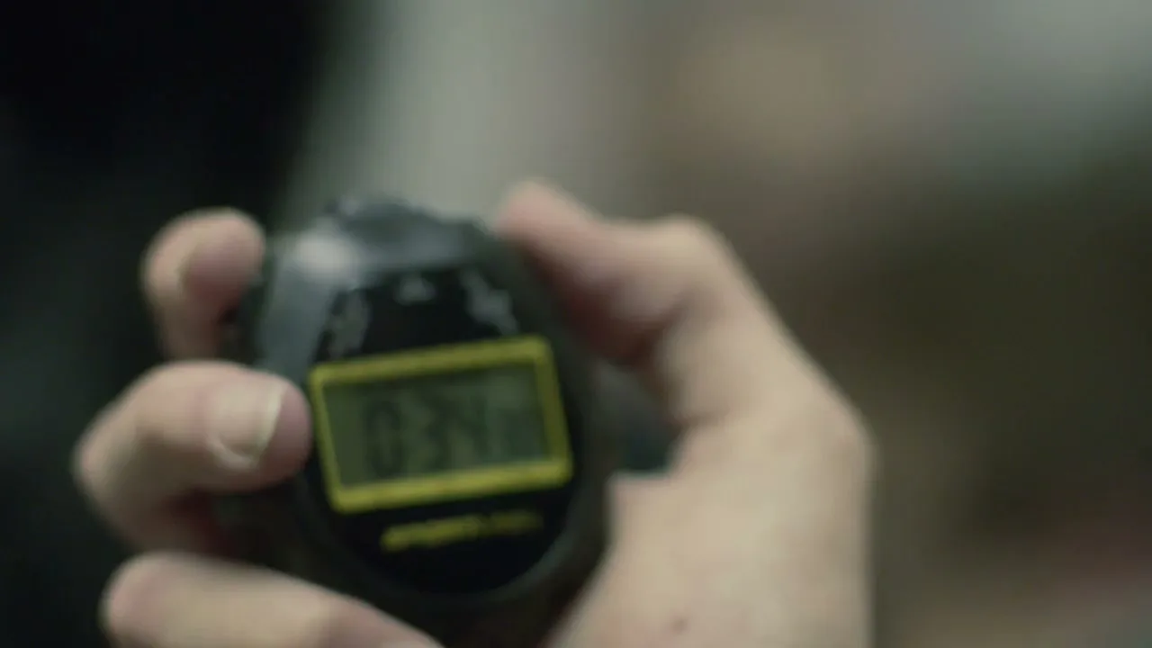 A person's hand holds a black digital stopwatch displaying "0:34:08" in a blurry, indeterminate indoor setting.