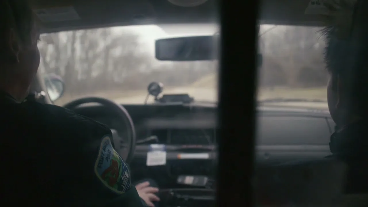 Two men, one in a police uniform, are seen from behind in the front seats of a police car, driving down a rural road on a cloudy day.