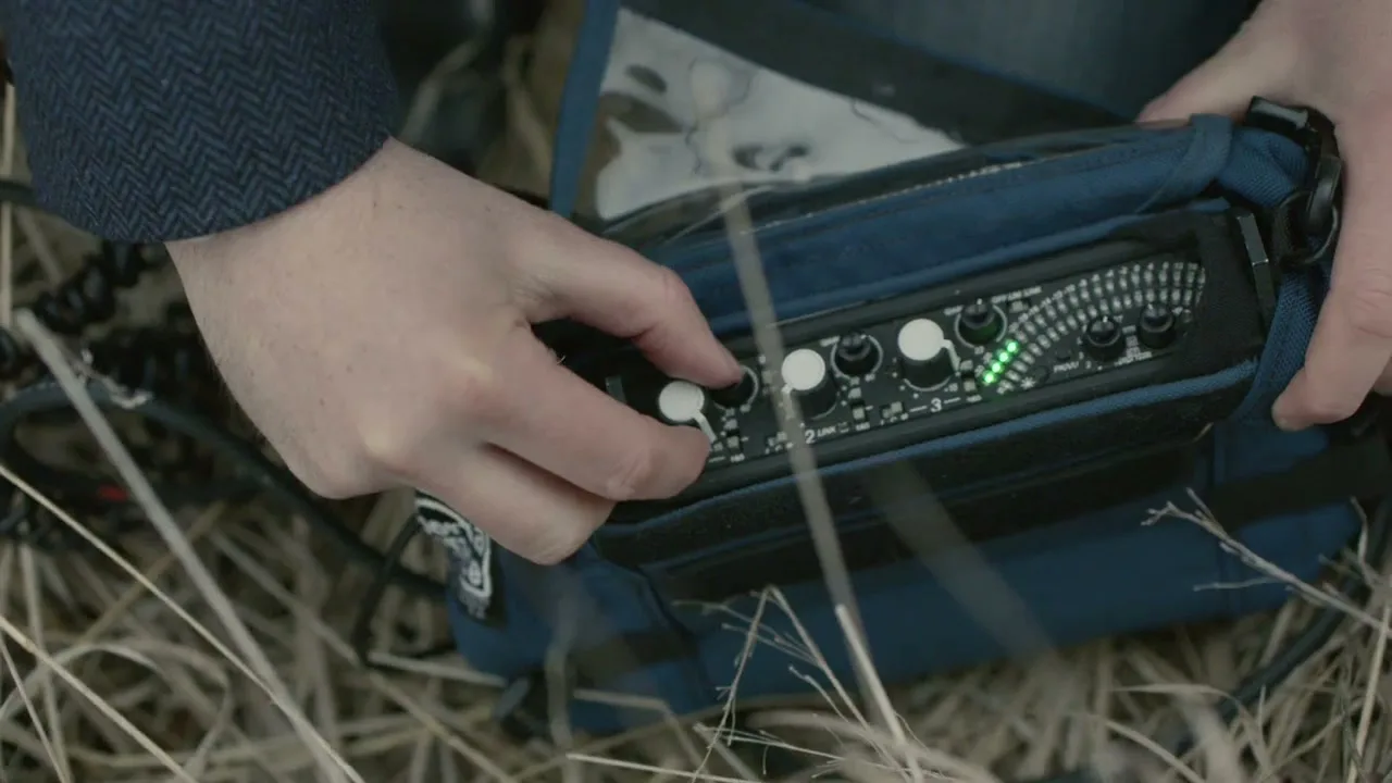 A person's hands adjust the knobs on a portable audio recorder, which sits in a blue case amidst dry grass.