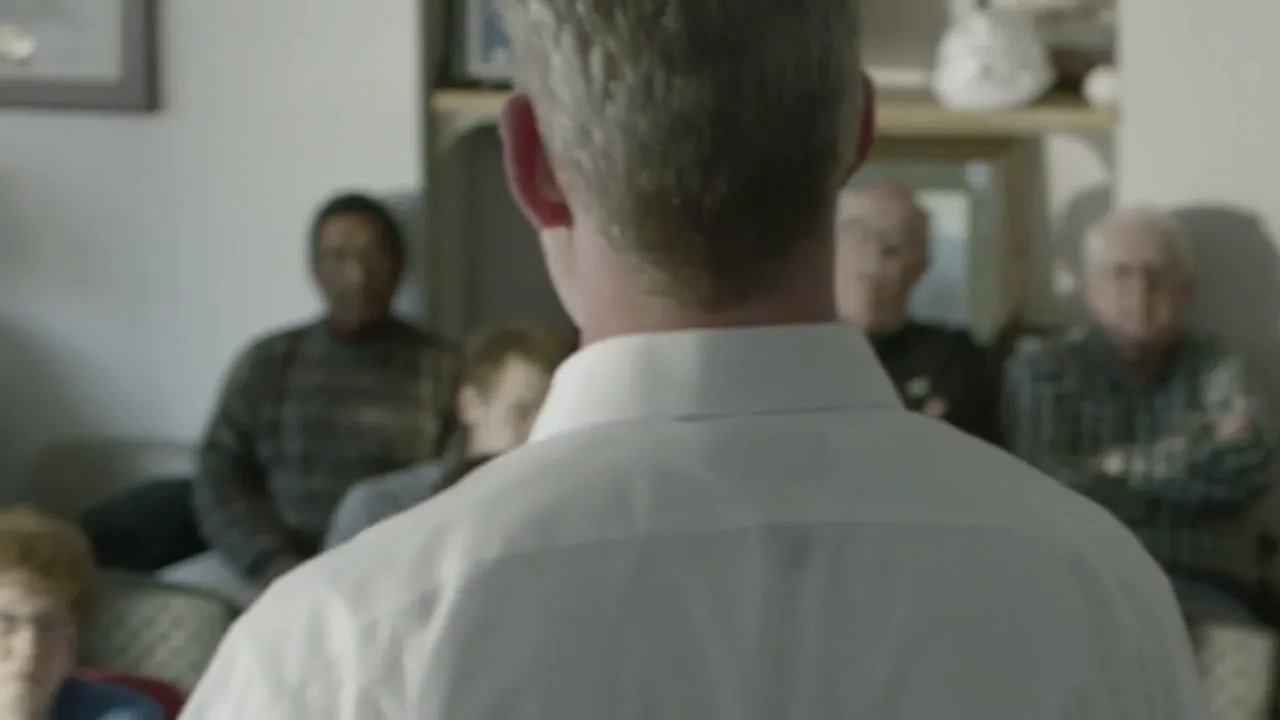 A man with gray hair and a white shirt stands with his back to the camera, addressing a diverse group of people seated in a living room.