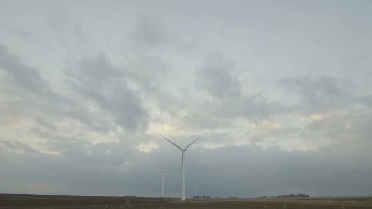 A wide shot of a vast, flat landscape with several wind turbines under a cloudy sky, with one turbine prominently in the foreground.