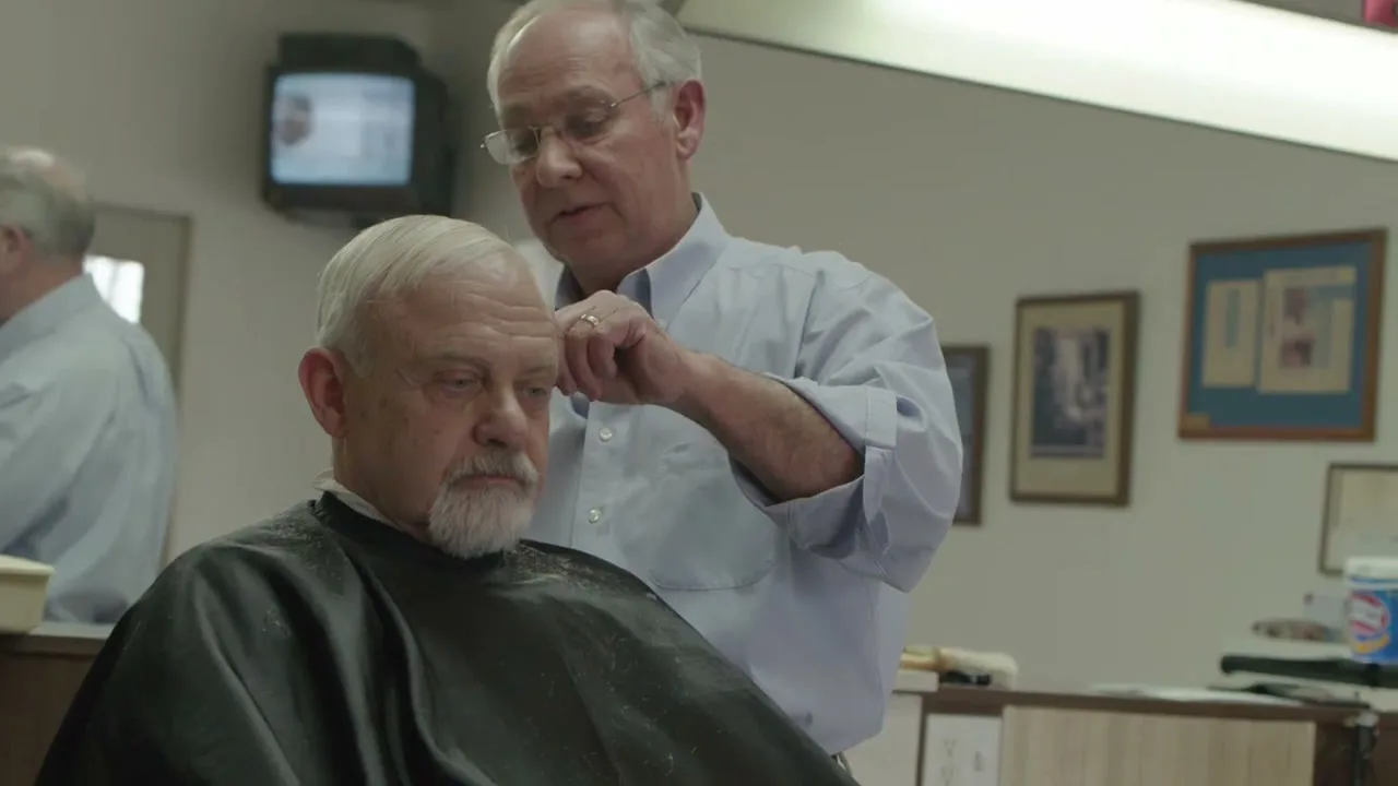 A male barber in a light blue shirt trims the hair of an older male client with a white beard, who is wearing a black cape, inside a barbershop.