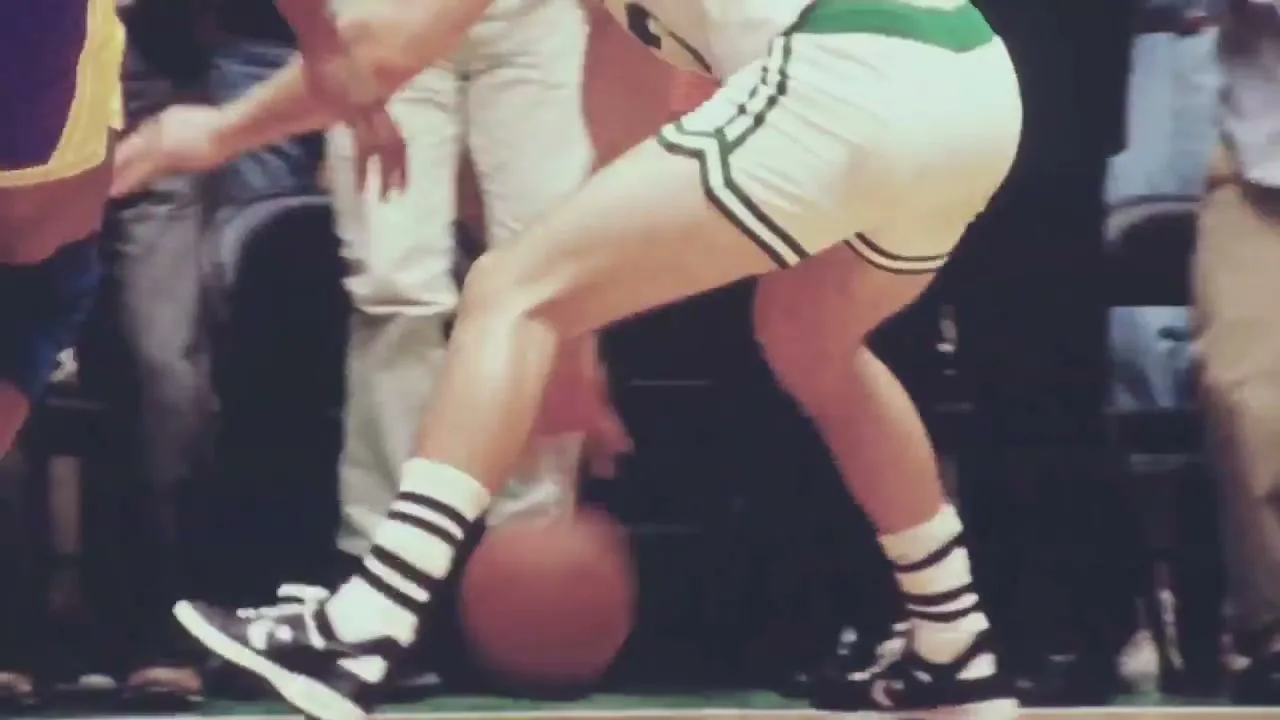 A low angle shot of basketball players' legs as they run and dribble on a court during a game, with spectators visible in the background.