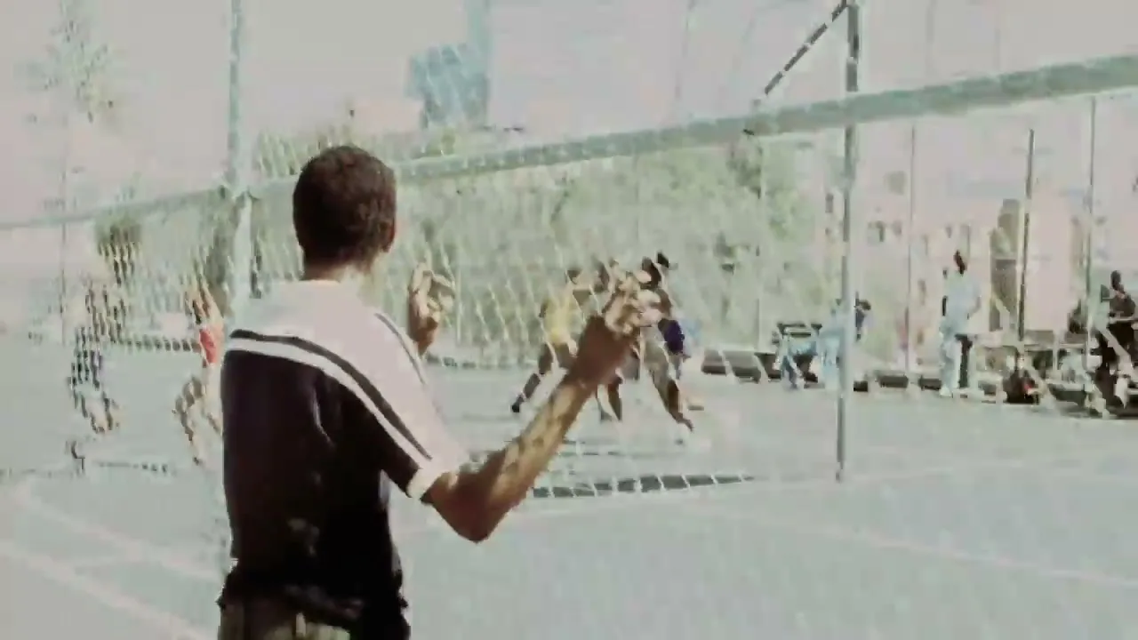 A young man in a black and white shirt watches a basketball game through a chain-link fence on an outdoor court during the day.