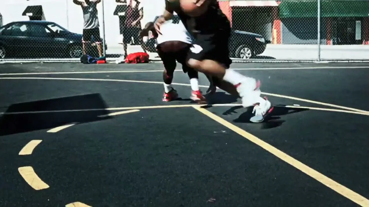 Two young men in athletic wear play basketball on an outdoor court, while other players watch in the background on a sunny day.