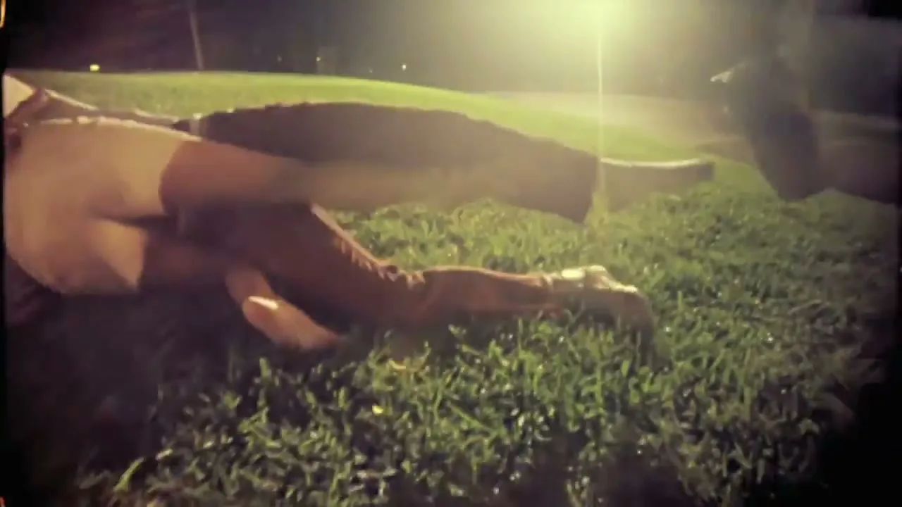 A person in brown pants and a white shirt is seen from a low angle, lying on green grass at night, with a bright light in the background.