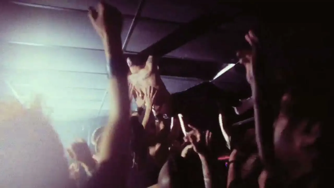 A shirtless man crowd-surfs at a concert, held up by many hands, under a dark ceiling with bright stage lights.