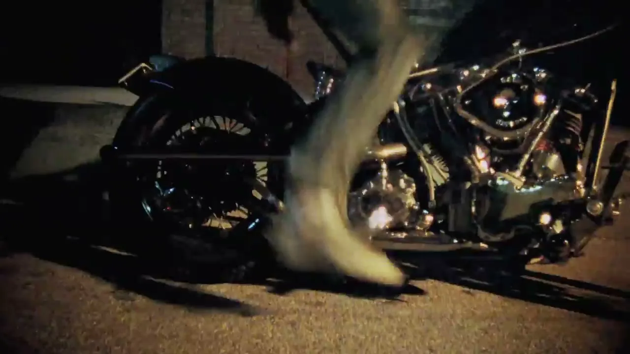 A person in jeans and white sneakers kicks the starter of a vintage motorcycle on a dark street at night.