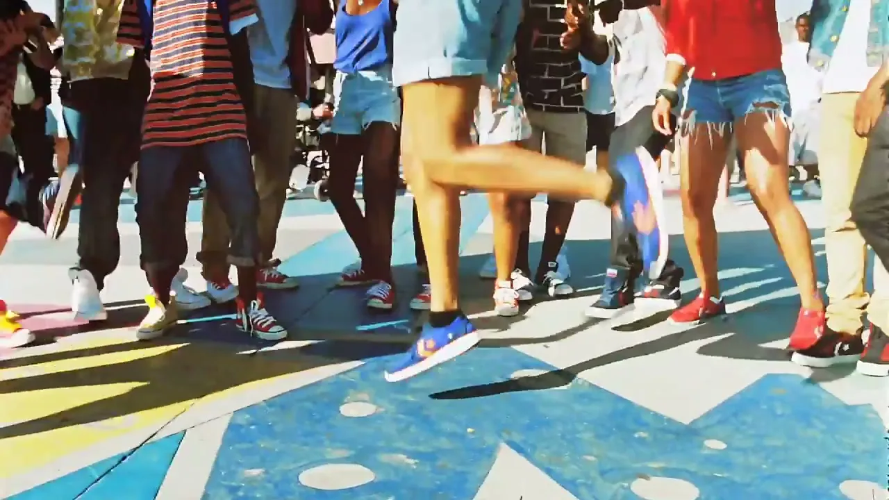 A diverse group of young people in casual attire and Converse sneakers dance energetically on a colorful outdoor pavement during the day.