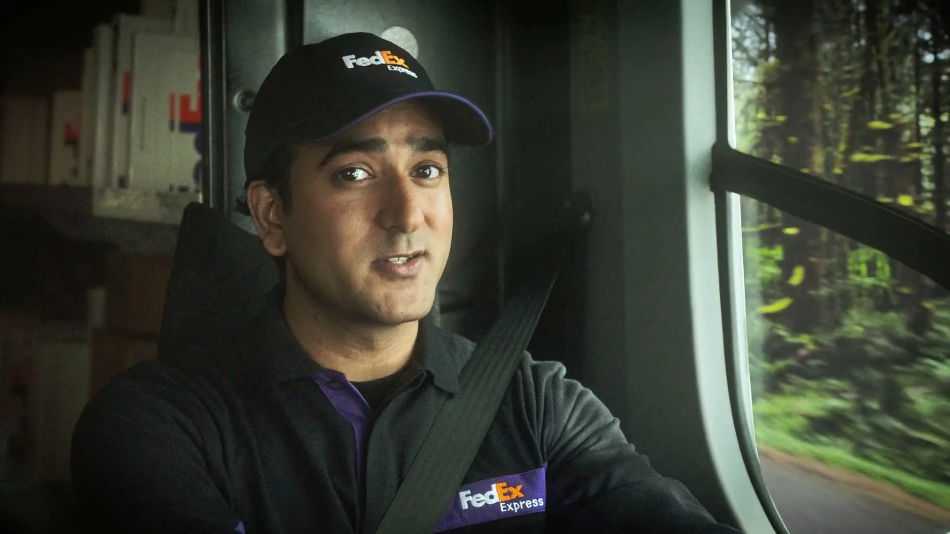 A male FedEx delivery driver in uniform, wearing a seatbelt, looks forward and speaks while driving through a forest.