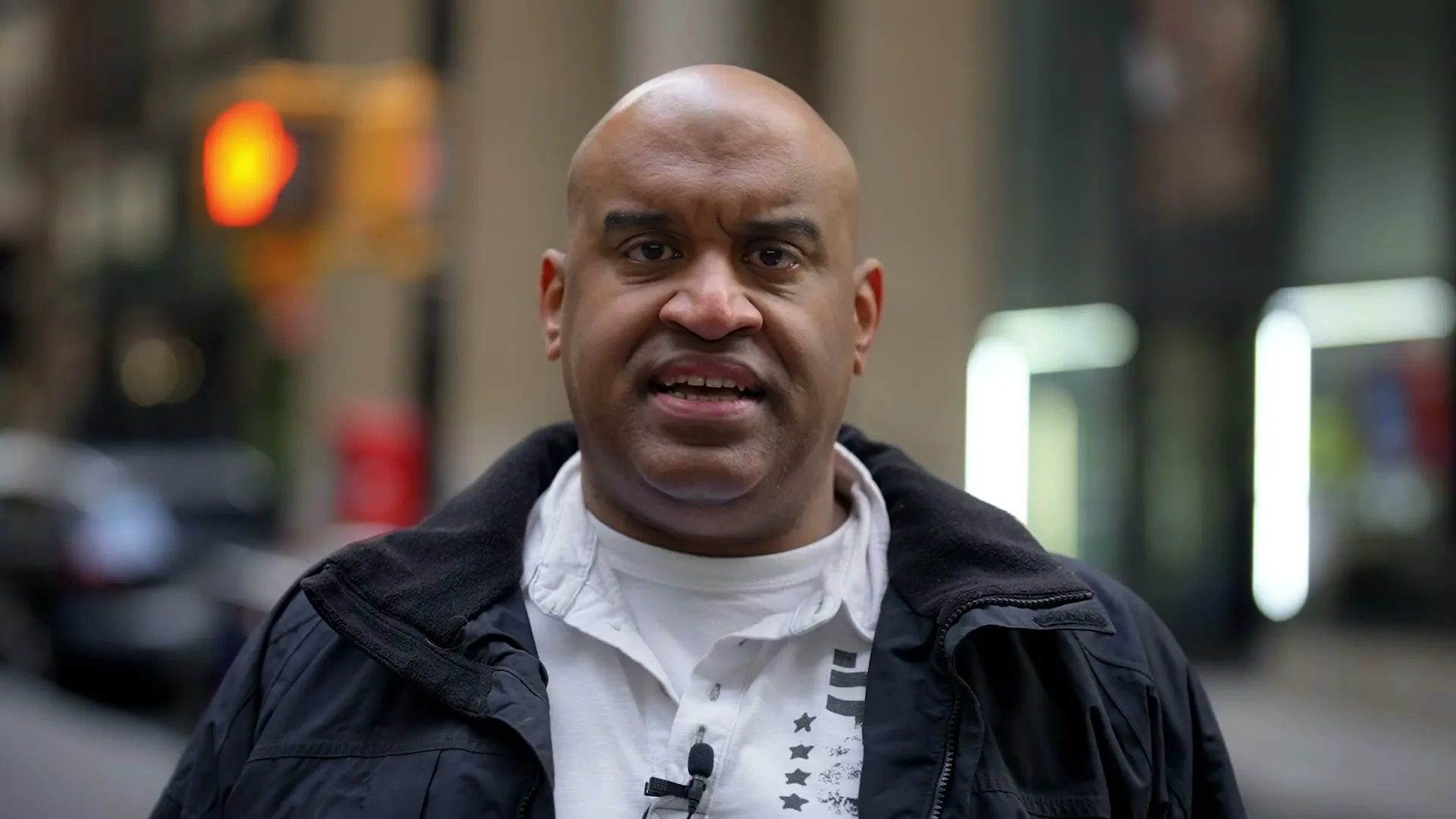 A bald man in his 40s, wearing a black jacket and white shirt, looks directly at the camera and speaks on a city street.