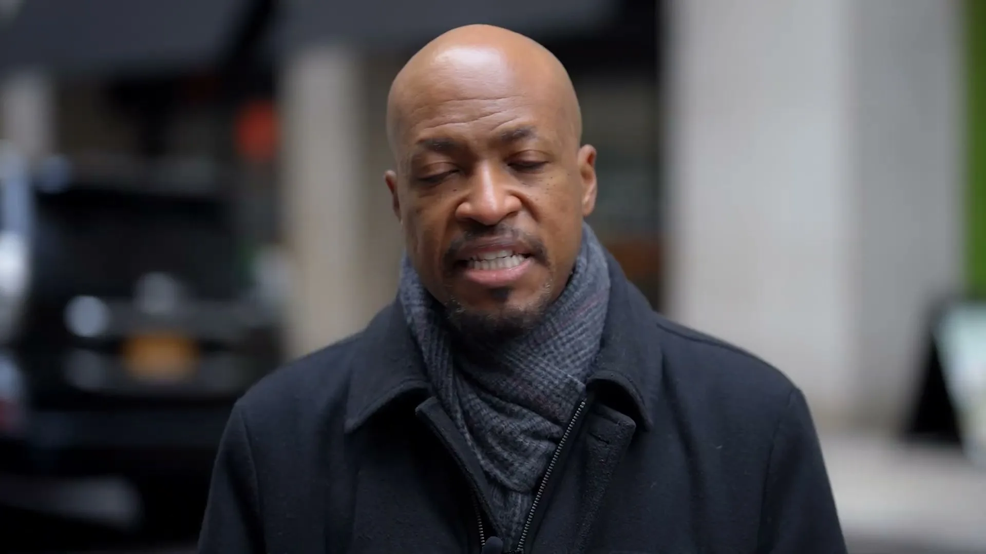 A bald Black man in his 40s, wearing a dark coat and scarf, looks directly at the camera with a concerned expression on a city street.