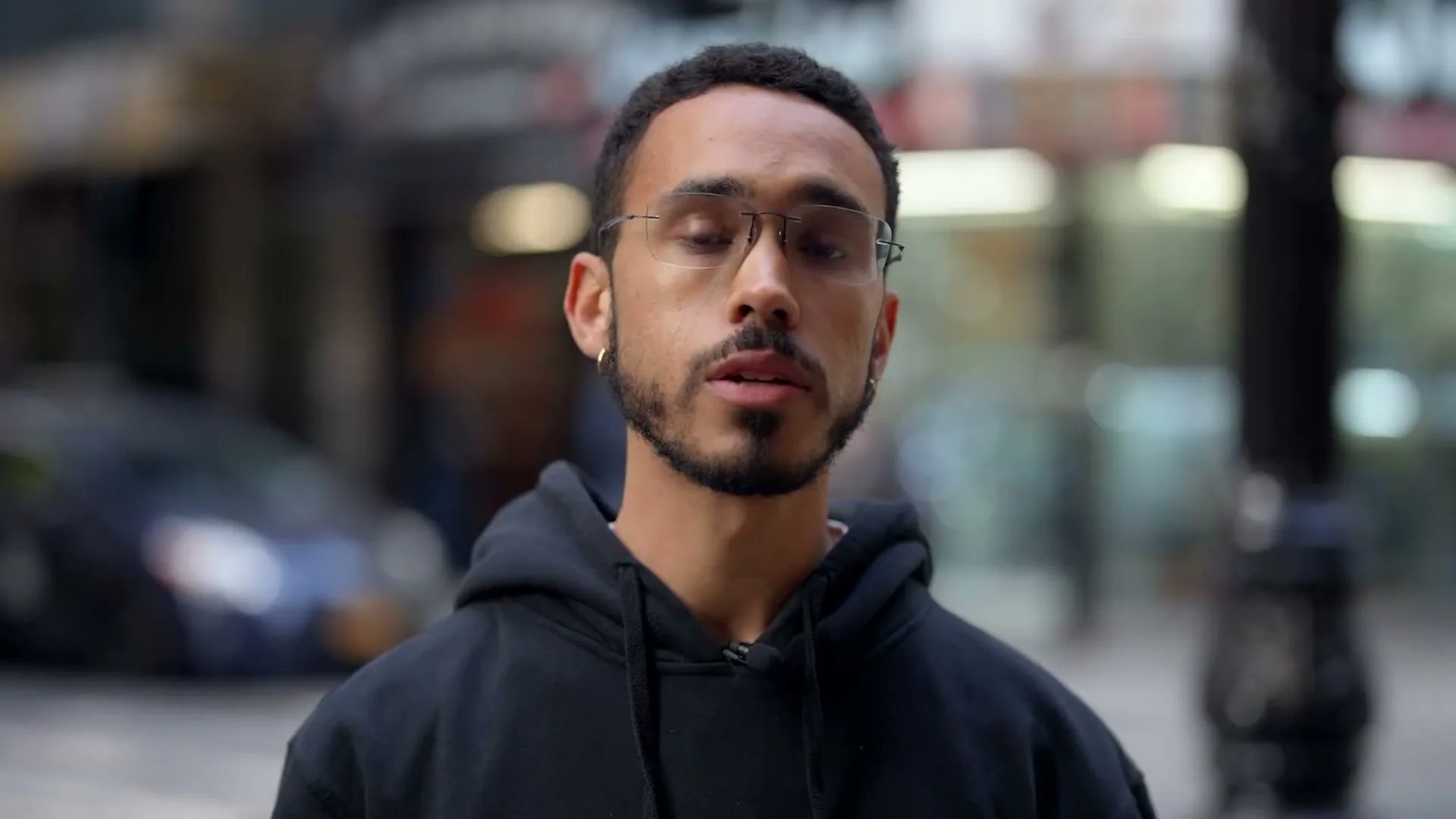 A young man with glasses and a beard opens his eyes and looks at the camera on a city street.