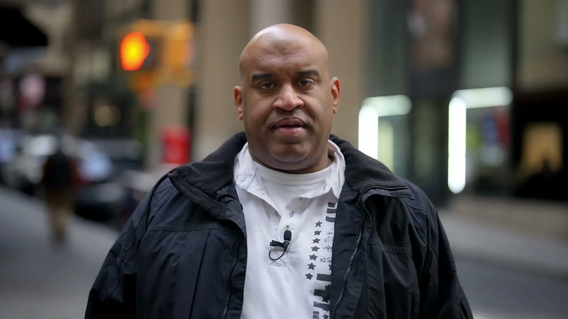 A bald Black man in his 40s, wearing a black jacket and white shirt, speaks directly to the camera on a city street.